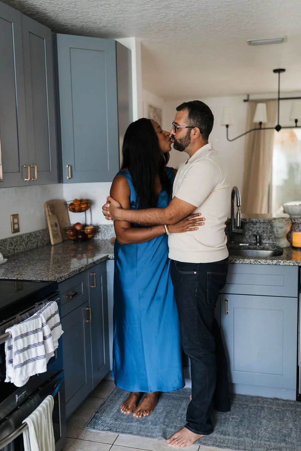 A couple sharing a kiss and embracing in a kitchen with blue cabinets, granite countertops, and a window with curtains.