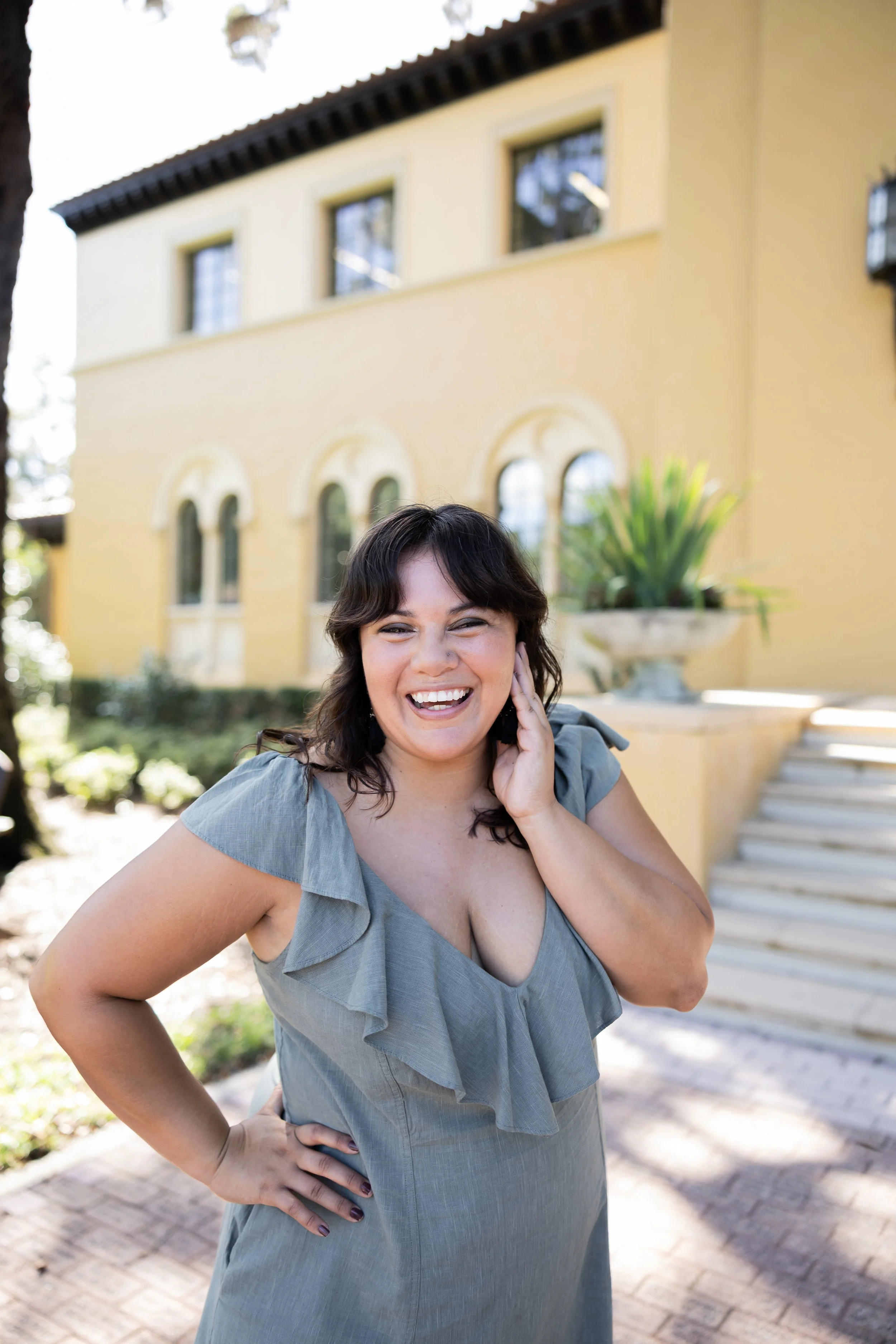 A woman with dark wavy hair smiling and touching her face, standing outdoors in front of a yellow house with arched windows and stairs, wearing a gray dress.
