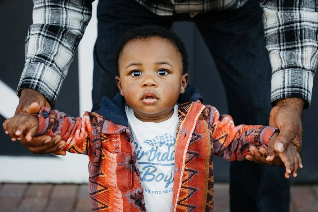 A young boy with dark skin and short curly hair receives support from an adult, holding his hands. The boy wears a colorful, patterned jacket and a birthday shirt, standing outdoors.