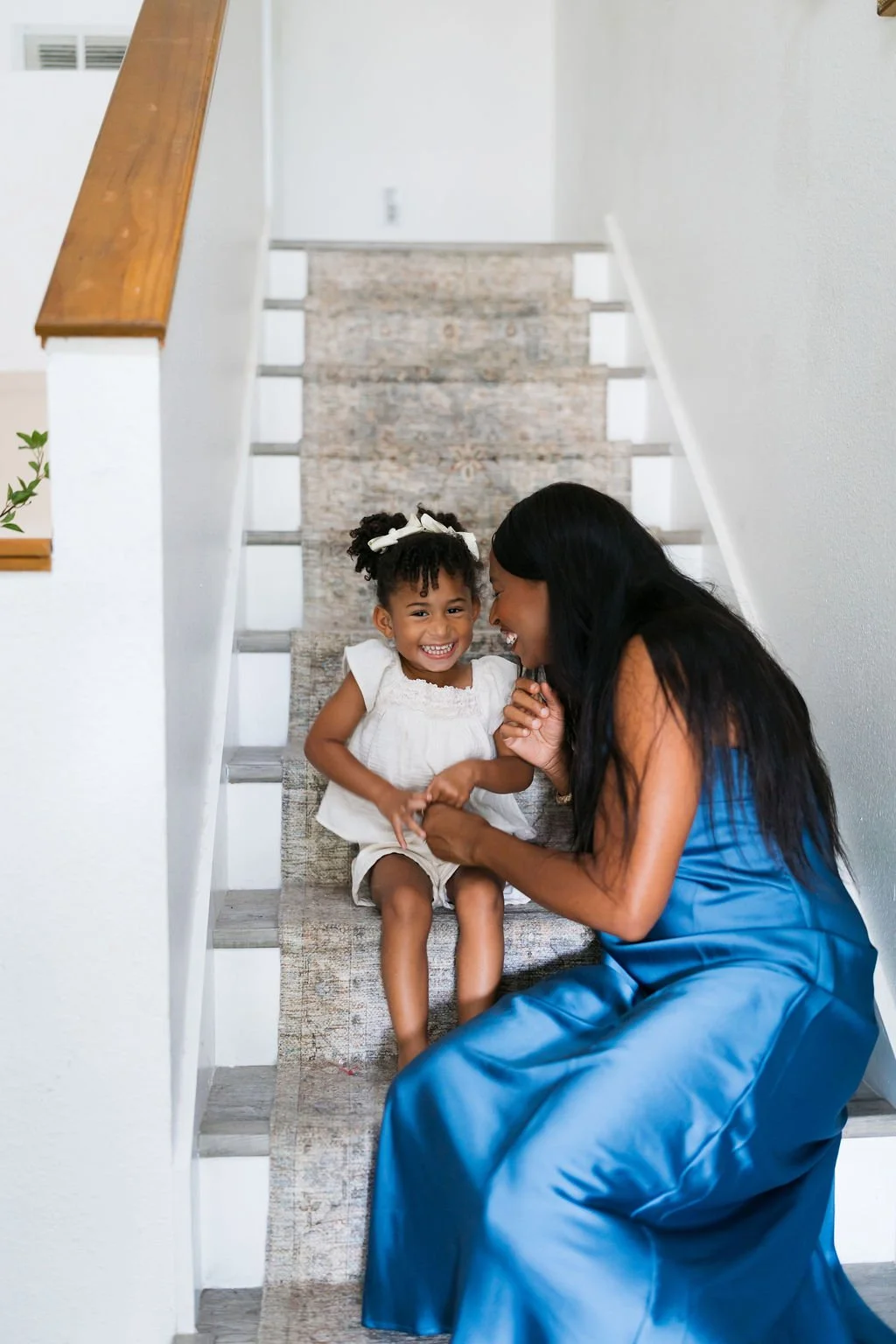 A woman and a young girl sit on a staircase, smiling and holding hands, sharing a joyful moment.
