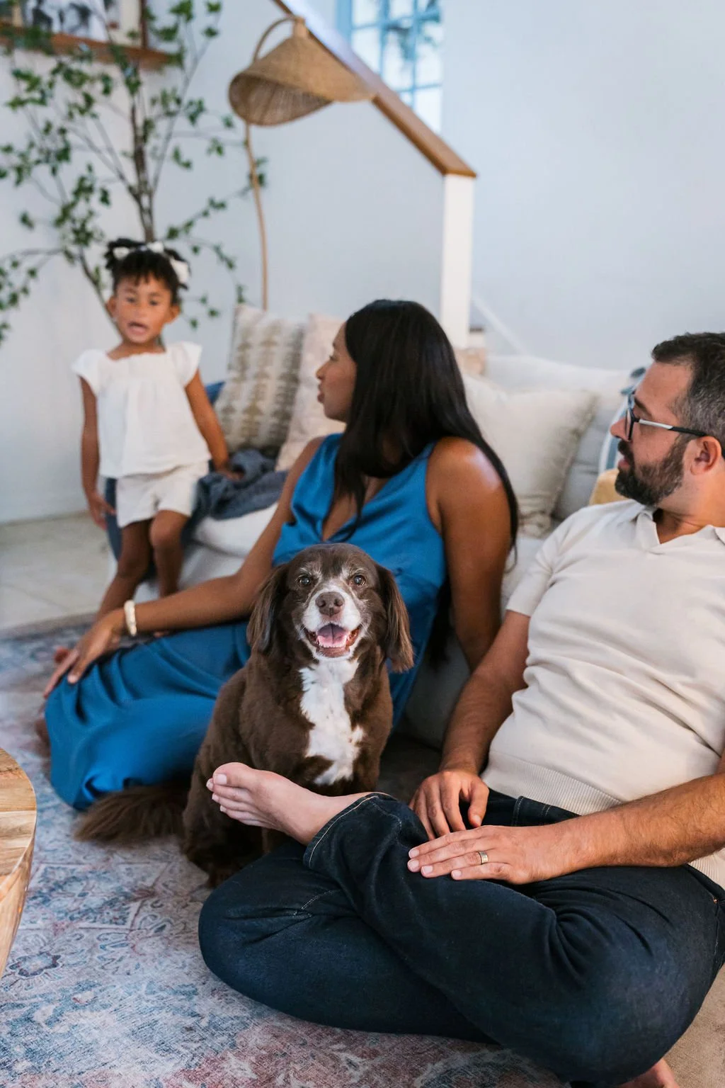 A family with a woman, man, young girl, and a brown and white dog sitting on a sofa in a living room, with the girl standing and the dog sitting close to the woman.