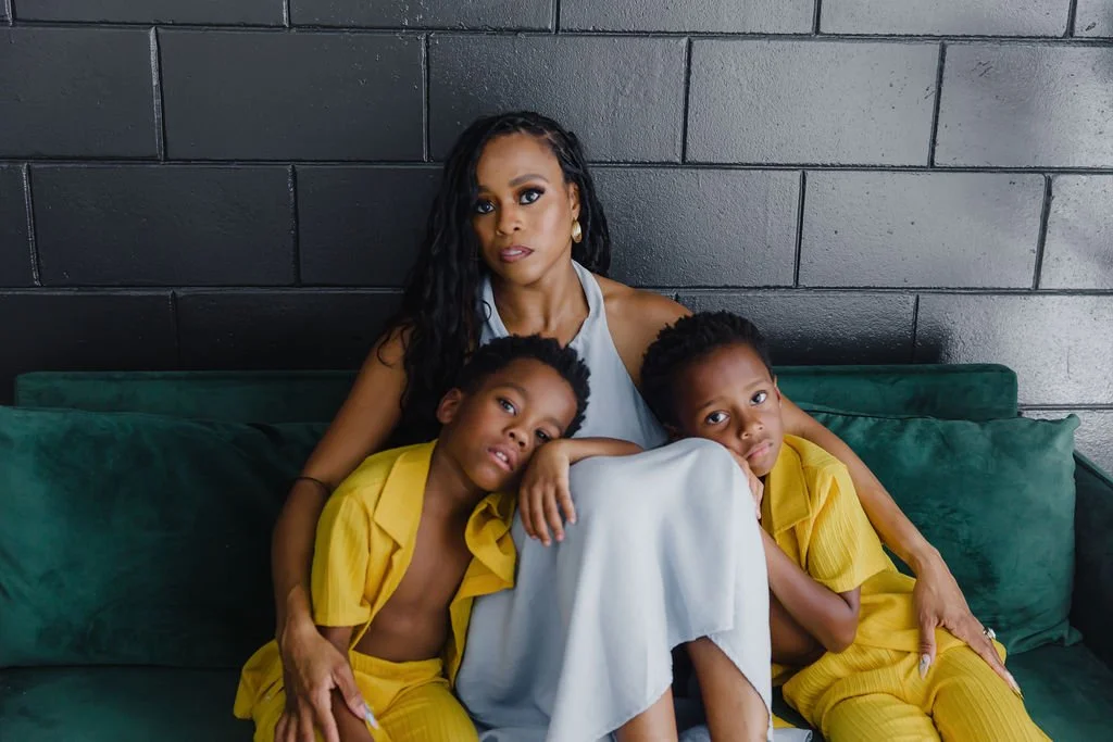 A woman and two young boys seated together on a green velvet couch against a black wall. The woman has long curly black hair and wears a sleeveless light-colored top. The boys wear yellow outfits, and all three have serious expressions.