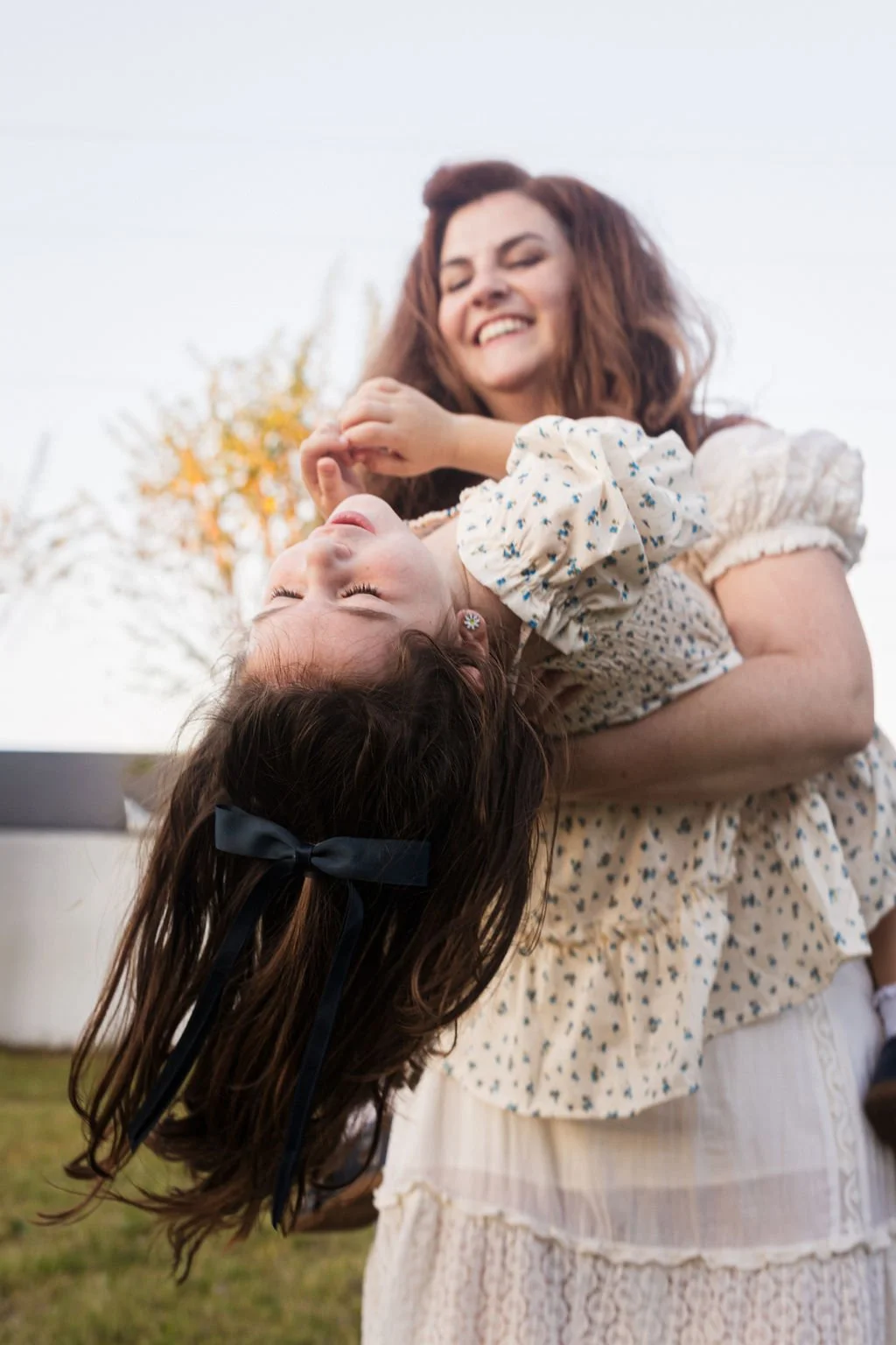 A woman holding a young girl in her arms, with the girl tilted backward and her eyes closed, outdoors with trees in the background.