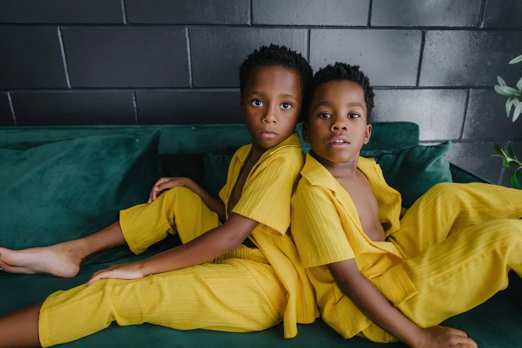 Two young boys sitting back to back on a dark green couch, wearing matching yellow pajamas, with a black brick wall behind them and a green plant on the right side.