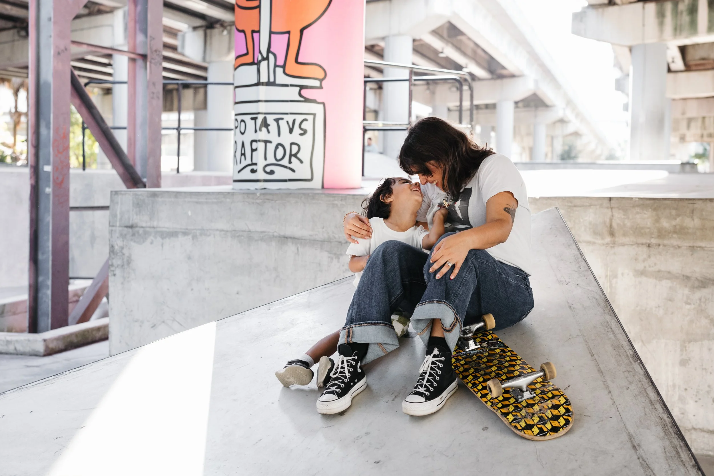 A woman and a young girl sitting on a skateboard at a skatepark under a bridge, sharing a joyful moment with their foreheads touching.