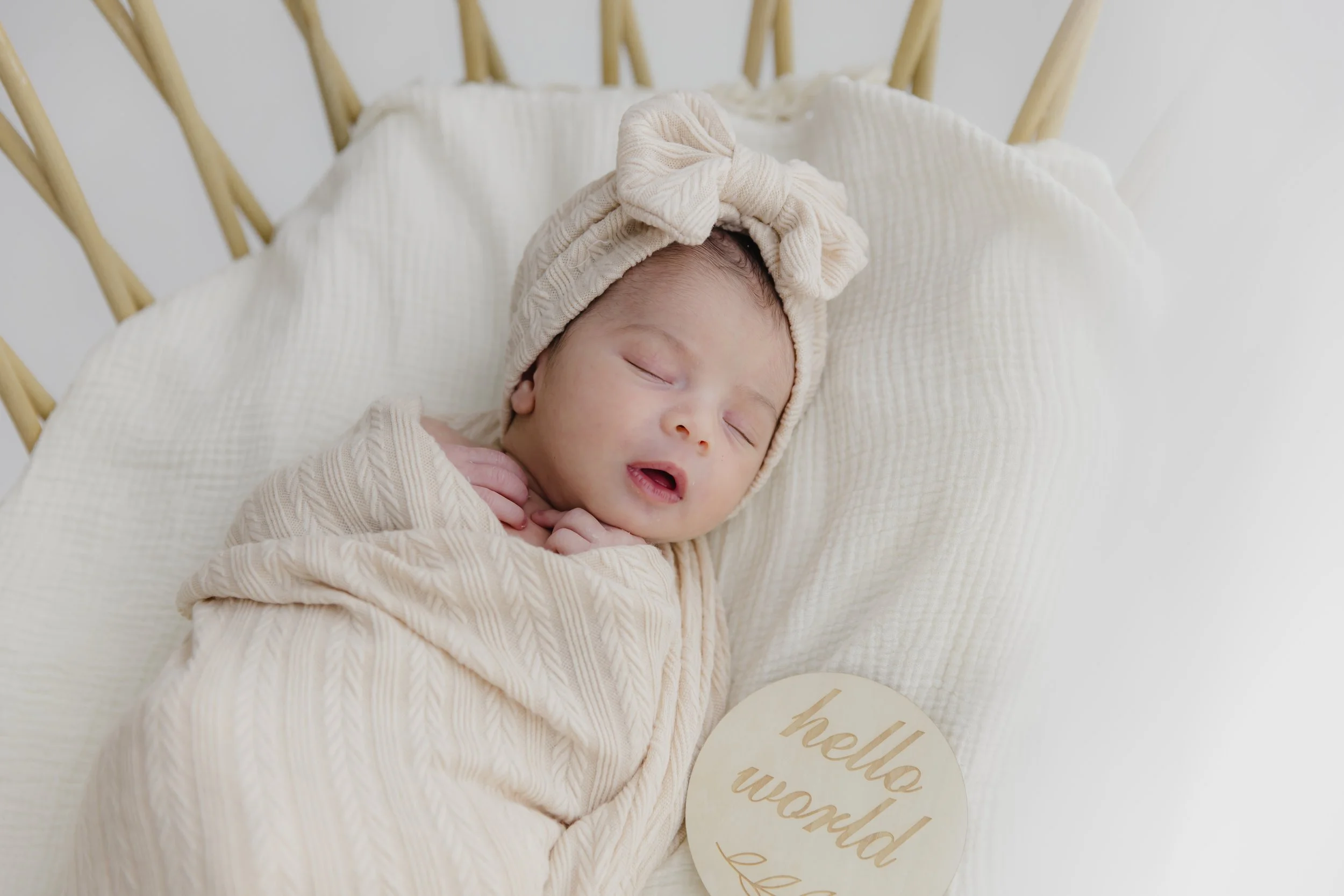 Sleeping baby wrapped in beige blanket with a matching headwrap, lying on a cream pillow, holding a round wooden sign that says 'hello world' in gold lettering.