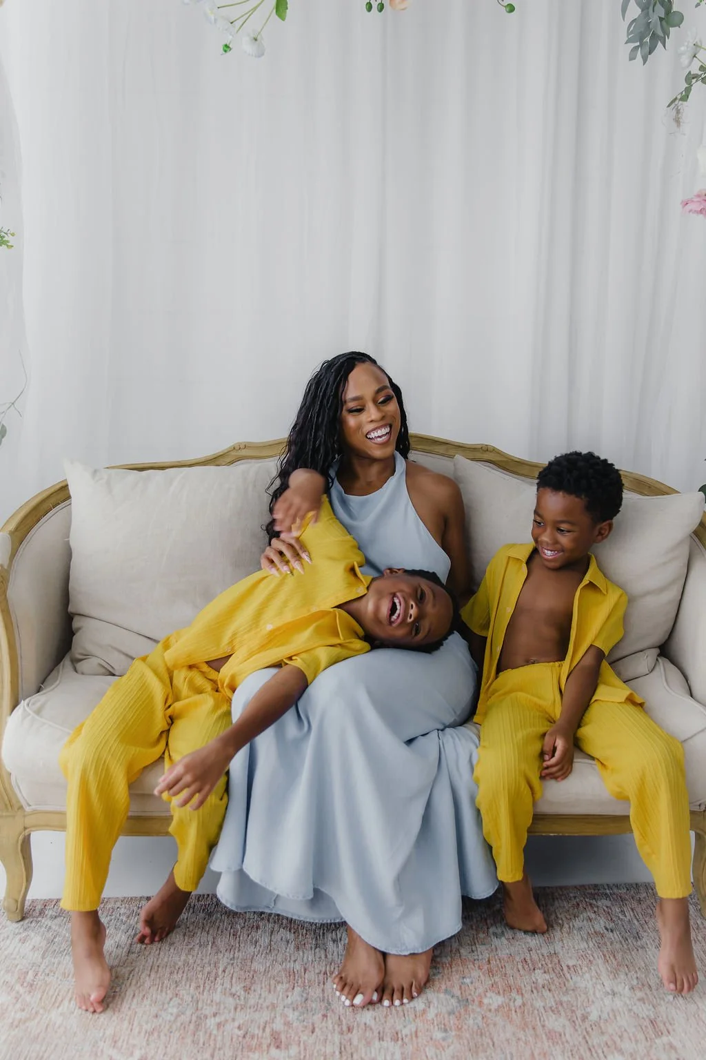 A woman and two young boys sitting on a cream-colored couch with a white backdrop and hanging plants. They are smiling and laughing, with the boys wearing matching yellow outfits.