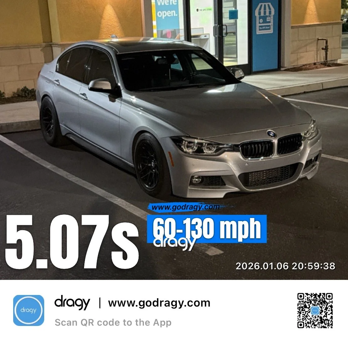 A silver BMW sedan parked in a parking lot at night outside a store with an "We're open" sign in the window.