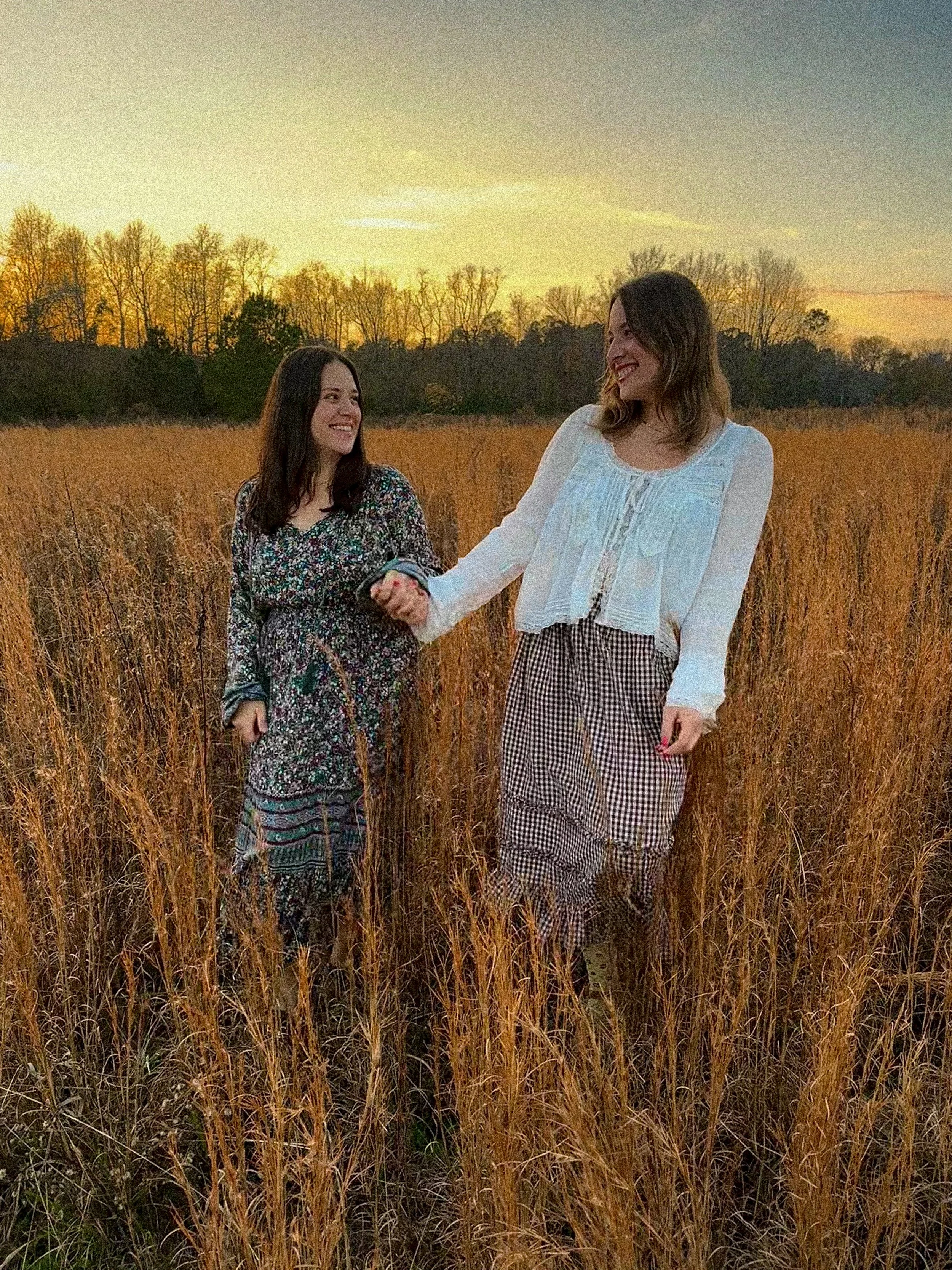 Two women hold hands and smile at each other in a field of tall grass during sunset.