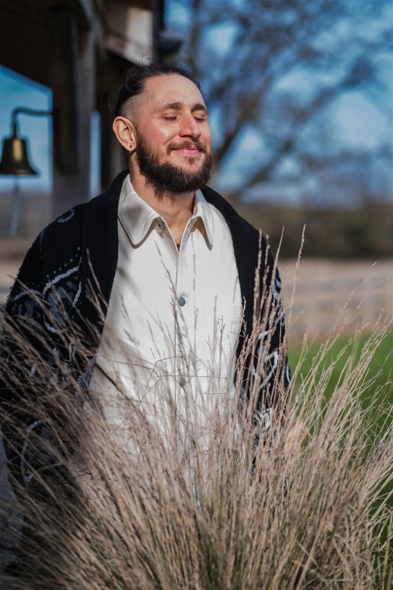 Dominic Hatcher, Internal Family Systems Therapist, standing outdoors near tall grasses, with a farmhouse and a tree in the background.