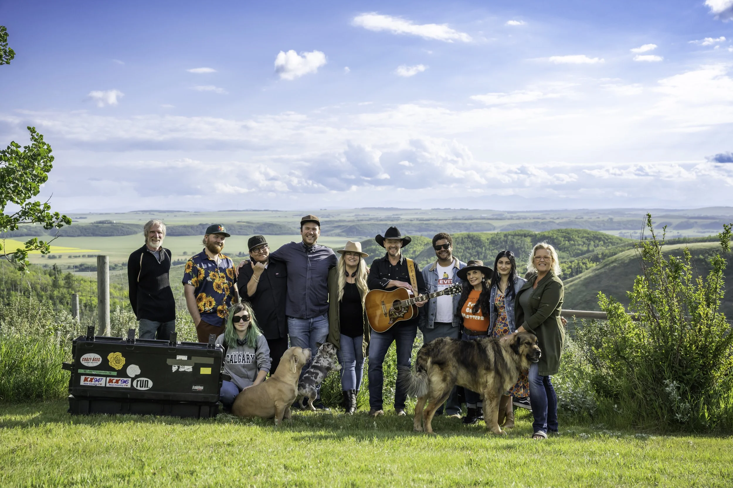 Scene from Not In My City, the crew photo outdoors in a green field, Paul Brandt holding his guitar centered with his wife, with dogs and a guitar case, under a partly cloudy sky.