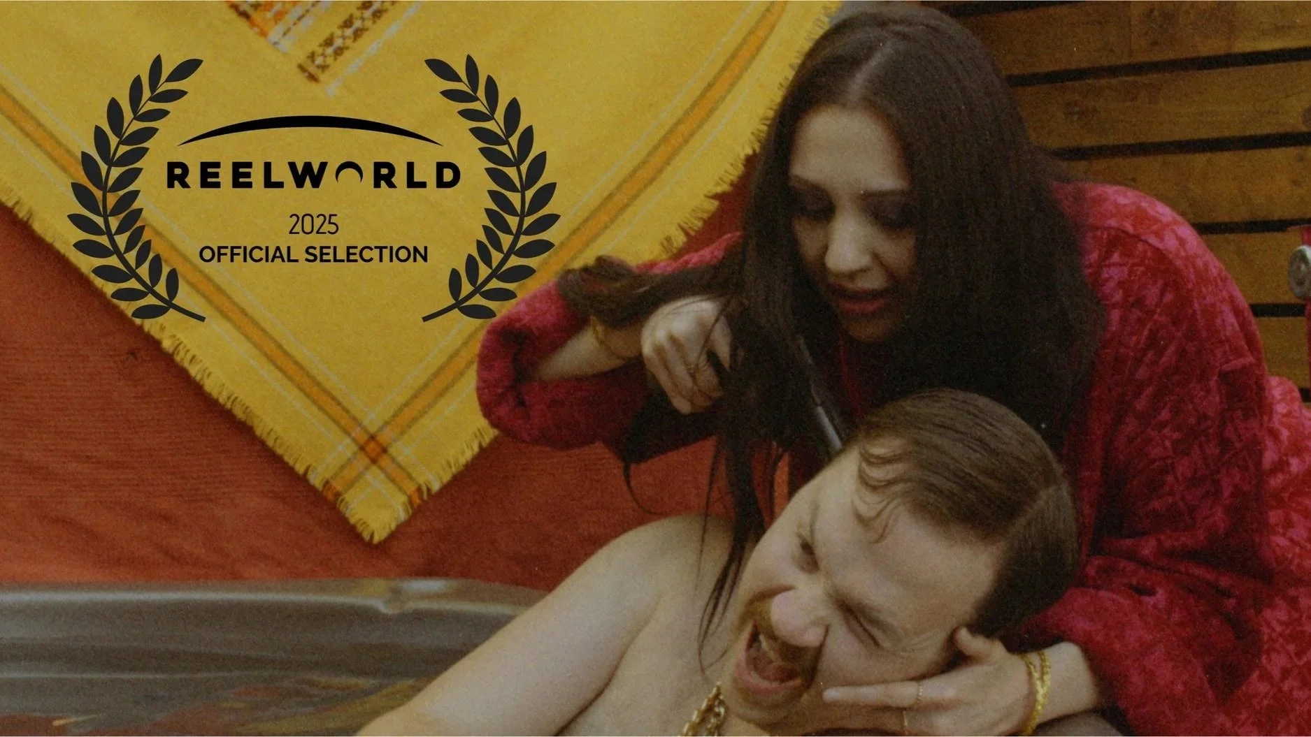 Scene from Renegade Fever showing a woman with dark hair in a red jacket holding a younger man with light brown hair and a gold chain at gunpoint, in front of a yellow cloth with a Reel World 2025 Official Selection logo.