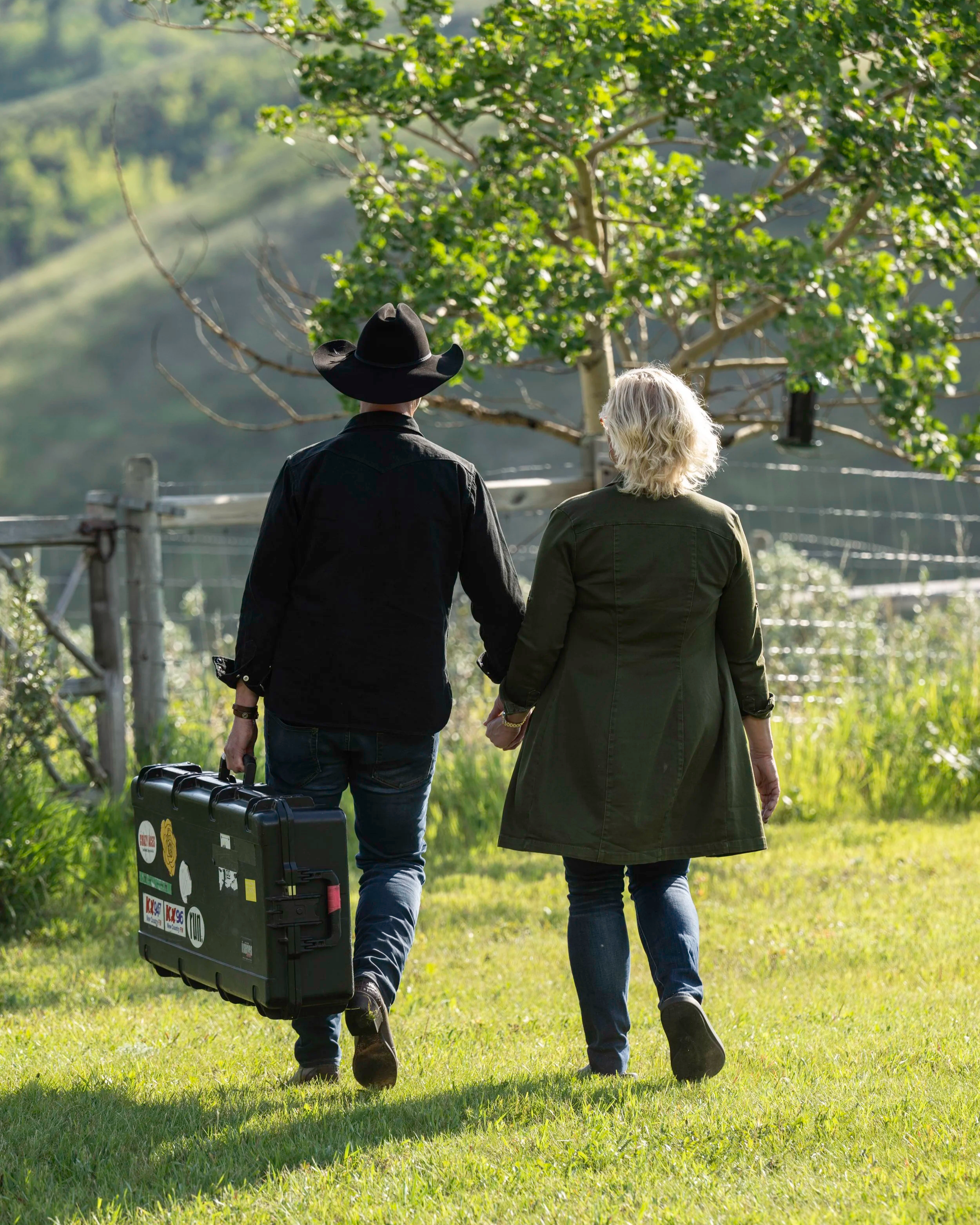 Scene from Not In My City showing Paul and Liz Brandt holding hands with their back to camera walking outdoors on a grassy area, with one carrying a black guitar case, surrounded by trees and greenery.