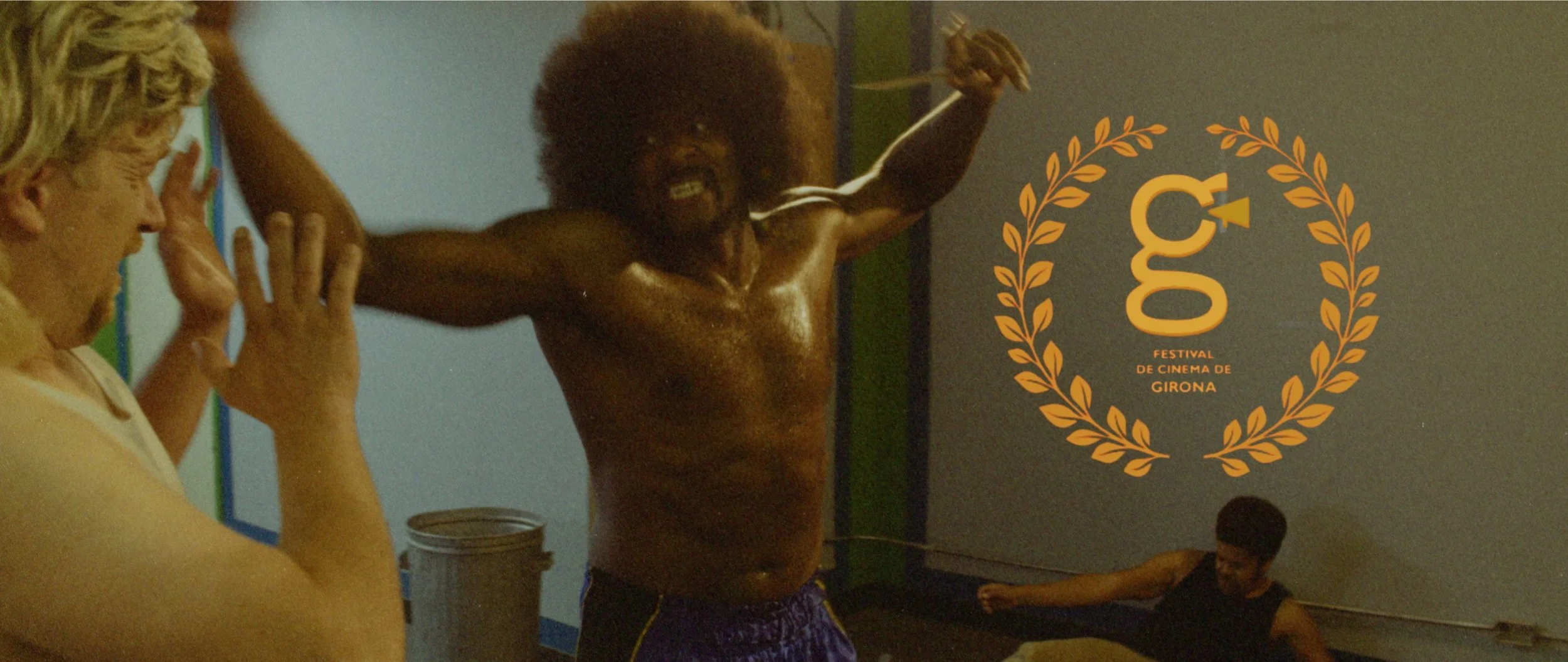 Scene from Renegade Fever showing a shirtless man with an afro hairstyle in a boxing stance, with a blonde man and another person in the background. The scene features a logo for the Girona Film Festival 2025.