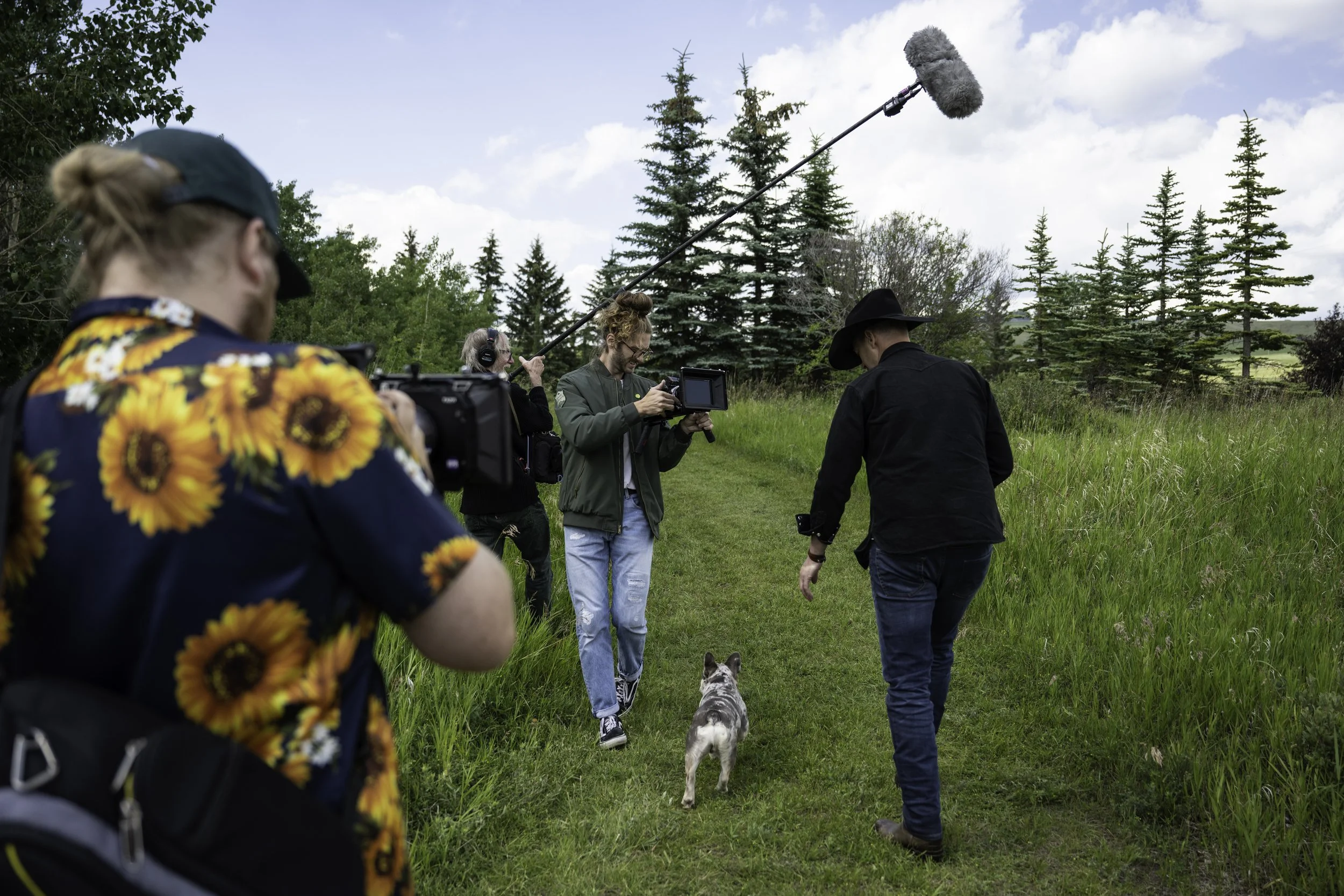 Scene from Not In My City filming crew members on a grassy trail with pine trees, capturing a scene where a Paul Brandt is walking with his dog, with a man holding a camera and a boom microphone operator recording above.