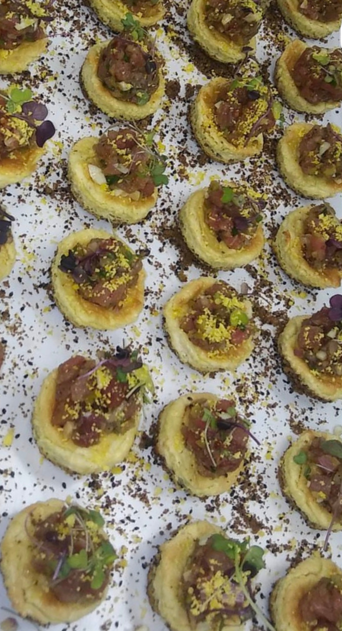 Mini toasts topped with a meat mixture, garnished with microgreens and yellow seasoning, arranged on a white surface with black and yellow crumbs.