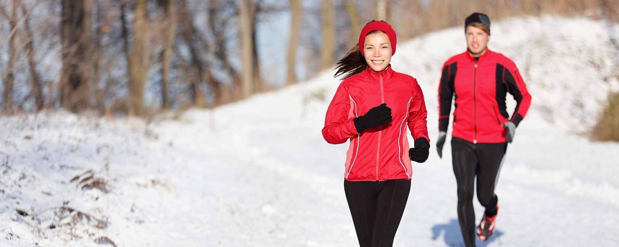 A woman and a man are running outdoors on a snowy trail during winter, both wearing red and black athletic jackets and black pants.