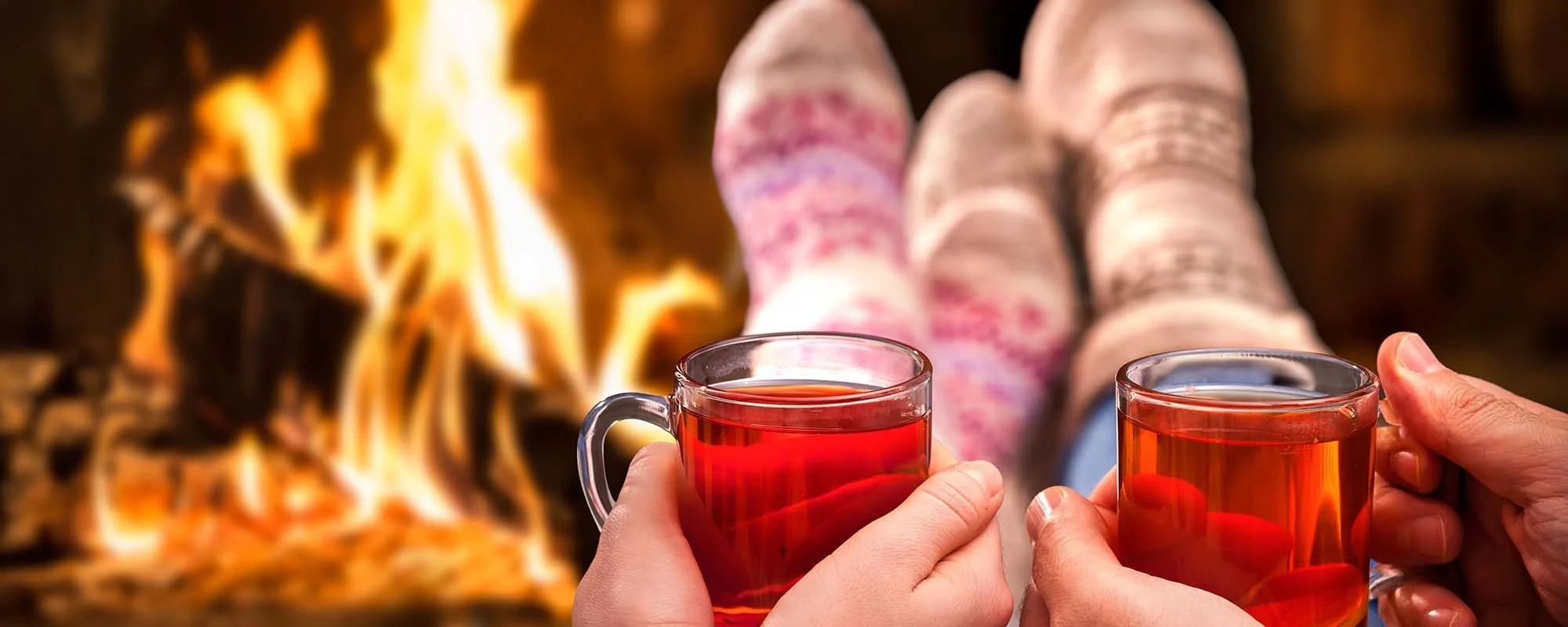 Two people holding clear glass mugs filled with a red beverage, relaxing in front of a lit fireplace, with their legs stretched out and wearing cozy socks.