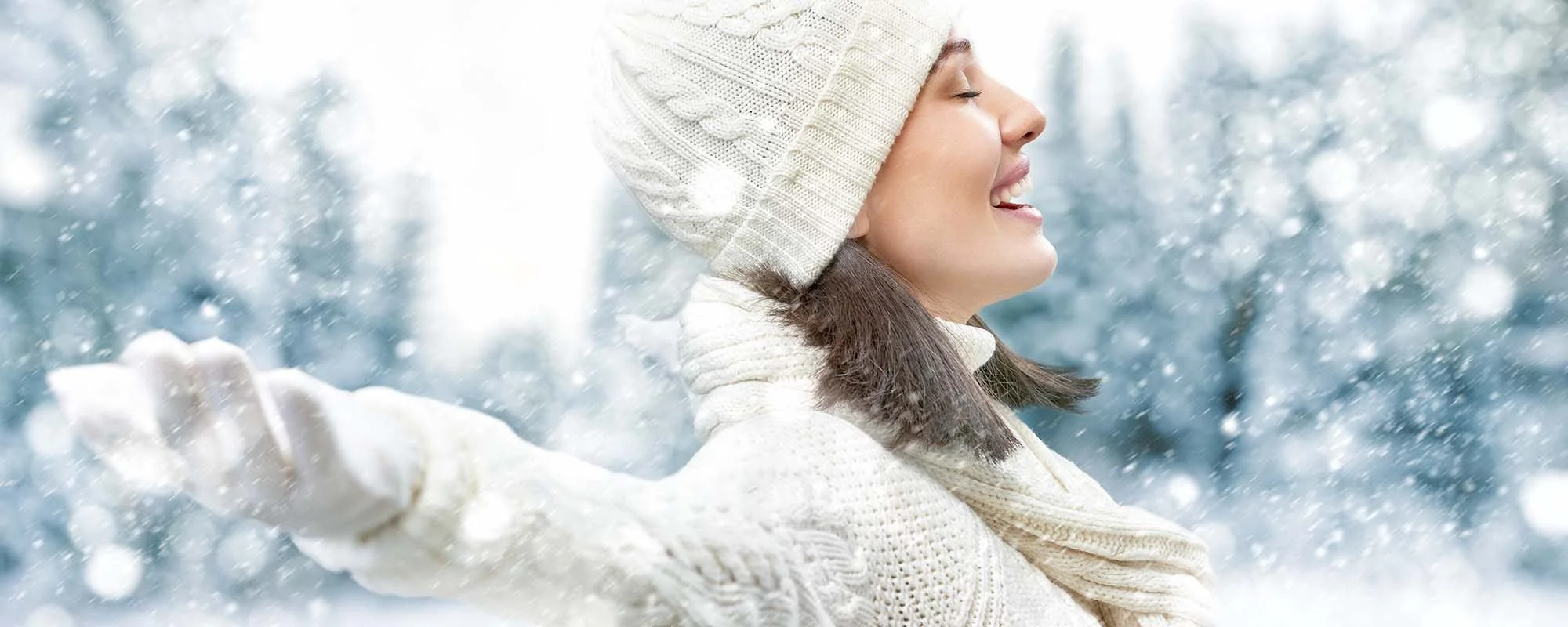 Woman in a wool hat and sweater enjoys snowfall with eyes closed and arms outstretched.