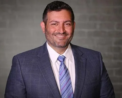 A man with dark hair and a beard, wearing a navy blue suit and a light blue striped tie, smiling in front of a gray brick wall.