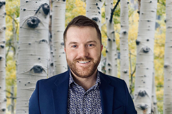 Jordan Dickson smiling in a professional headshot, wearing a navy blazer and patterned shirt, with a blurred tree background.