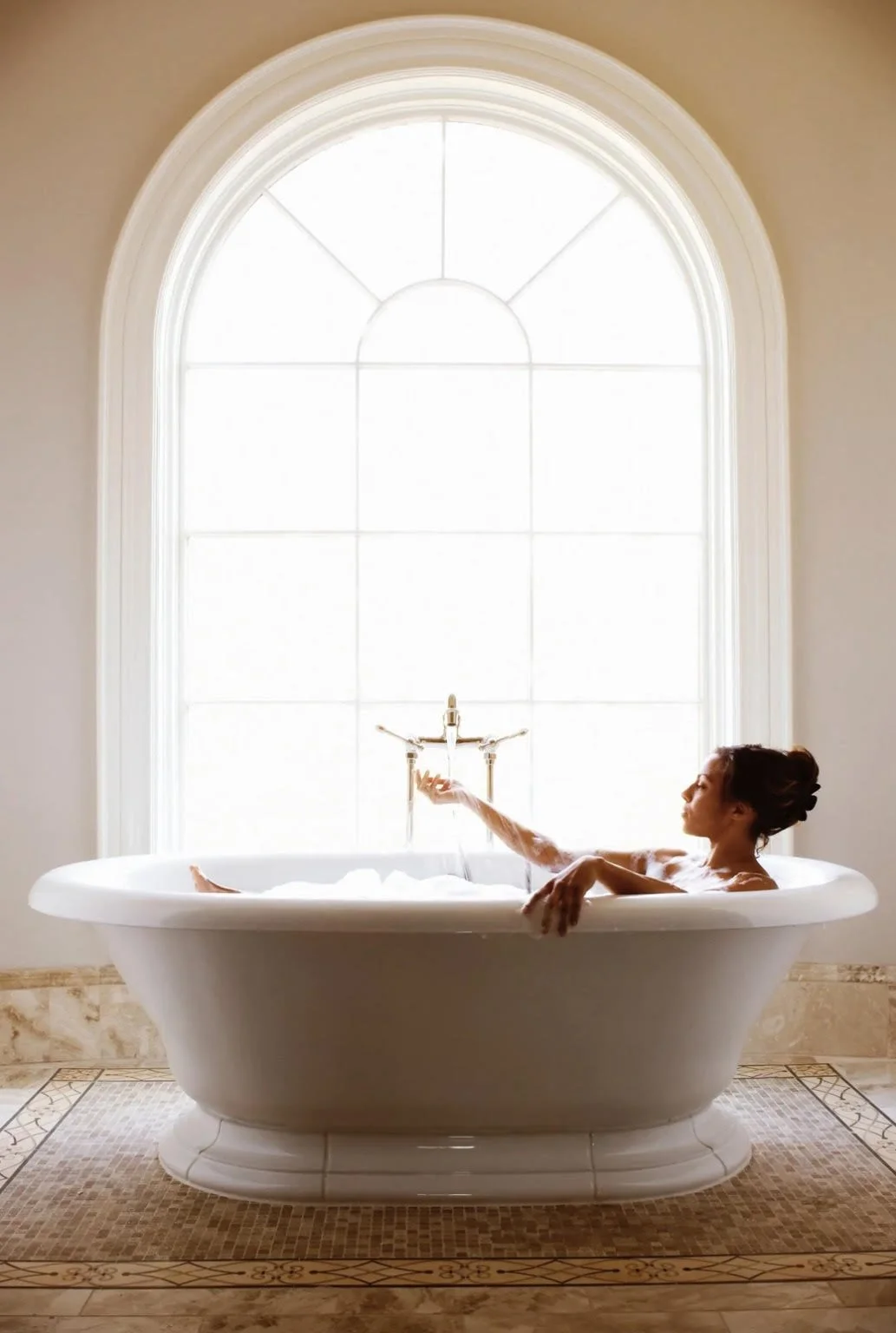 A woman relaxing in a bathtub near a large arched window with bright sunlight streaming in.