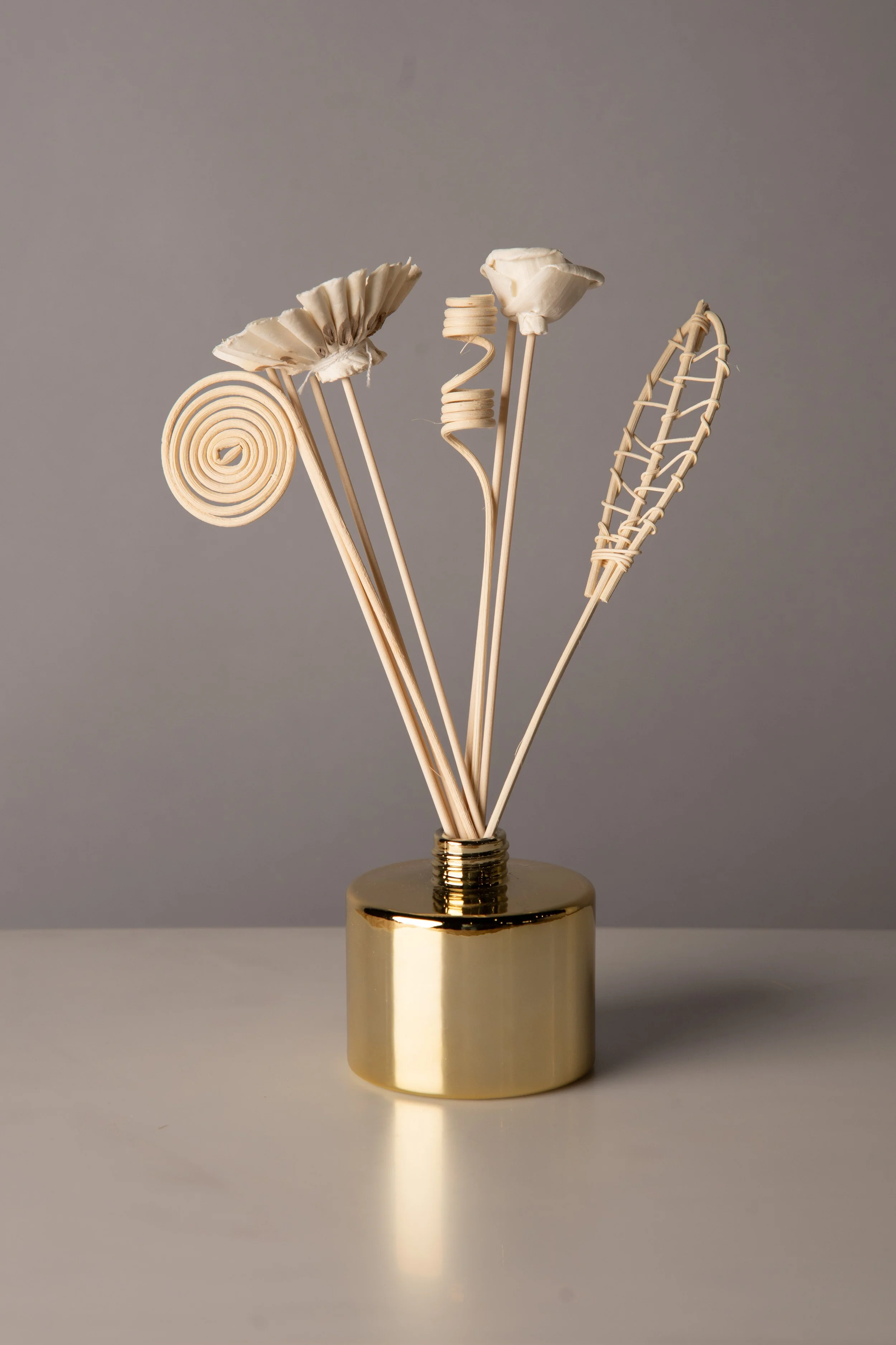 A gold-colored container holding three decorative reed sticks with different shapes: one spiral, one flat and woven, and one simple straight stick.
