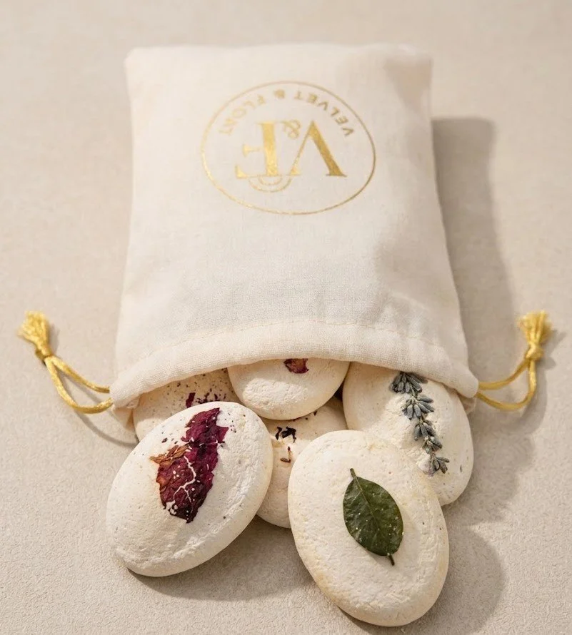 Close-up of botanical Shower Pebbles beside the ivory Velvet & Float drawstring pouch. Assorted styles shown; the bundle contains six in your chosen fragrance.