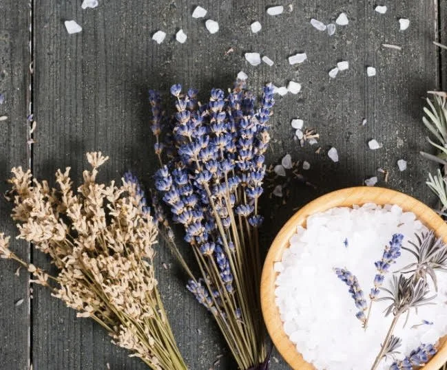 Baskets of dried lavender and other herbs on a dark wooden surface with scattered white flower petals.