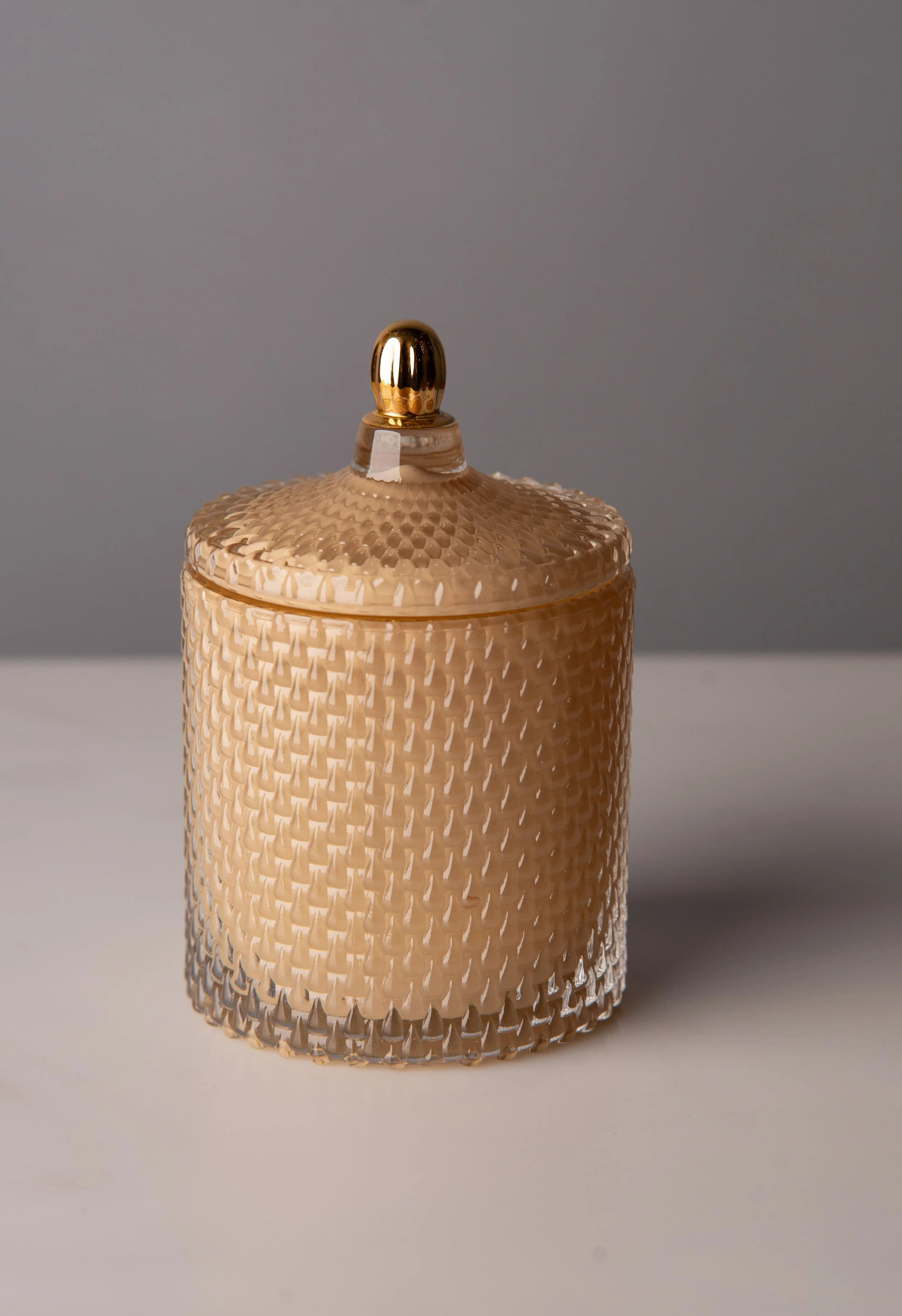 A gold decorative jar with a textured surface and a lid, accompanied by a matching small scoop or spatula, set against a plain beige background.