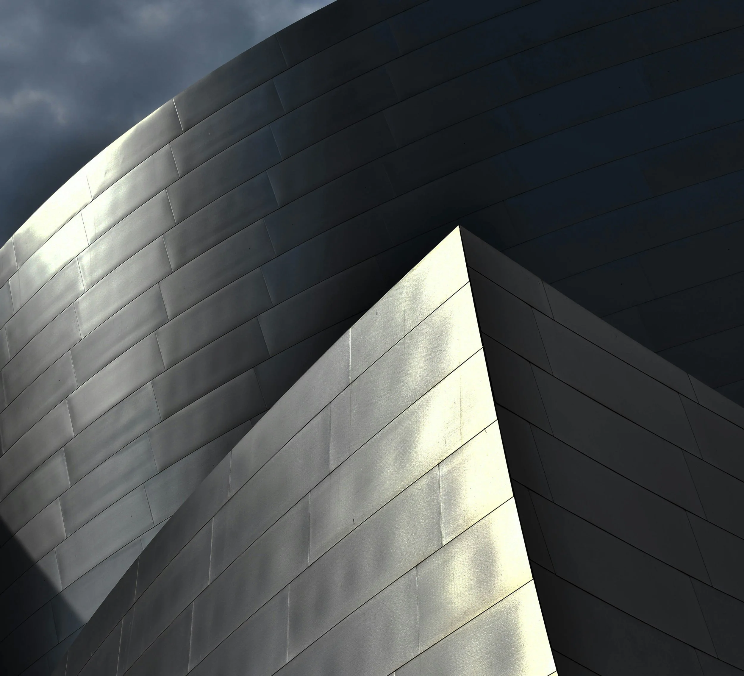 Close-up of a modern building with metallic gray panels, featuring intersecting curved and angular surfaces, under a dark sky.
