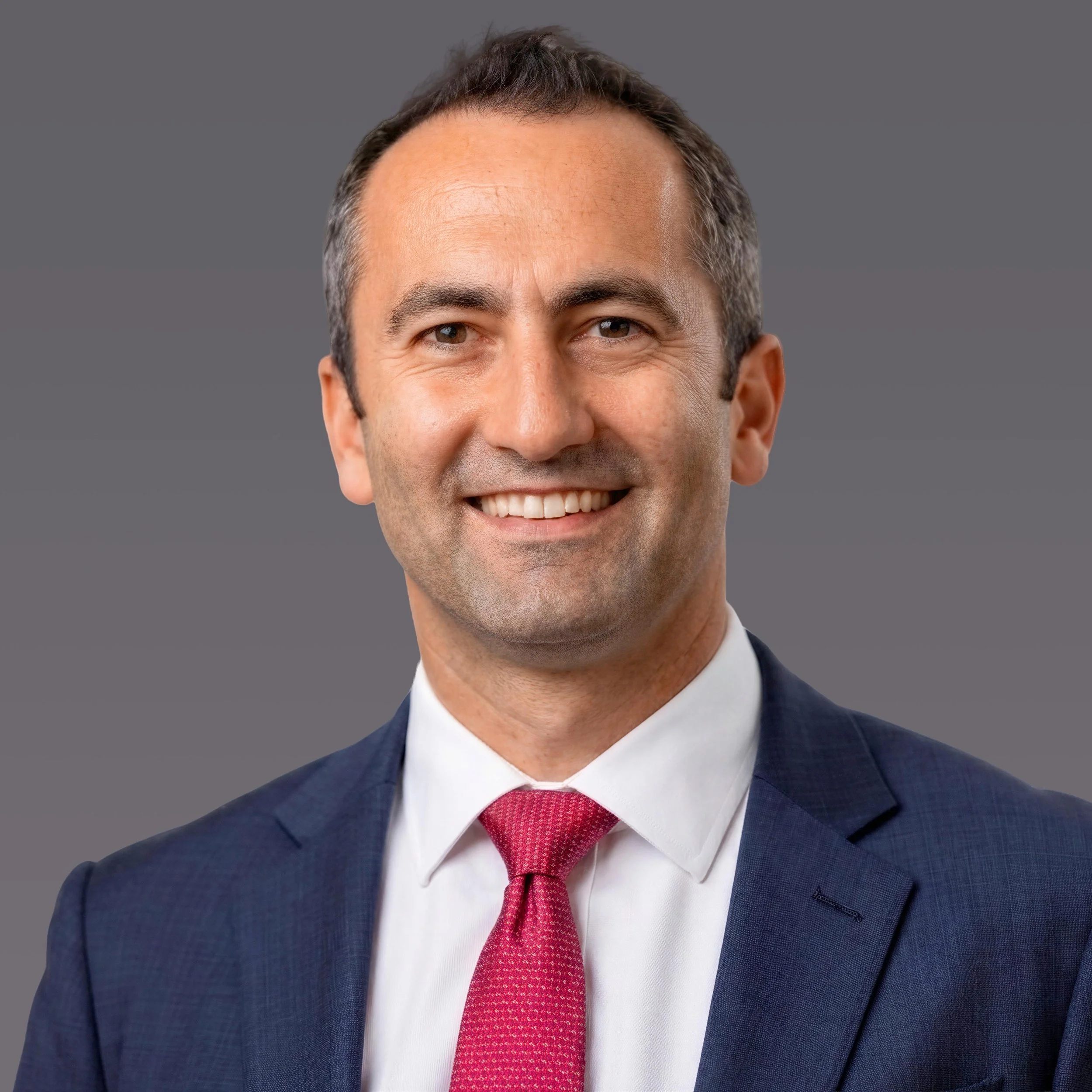 Headshot of a Matthew Jacobs smiling in a navy suit, white shirt, and red tie against a gray background.