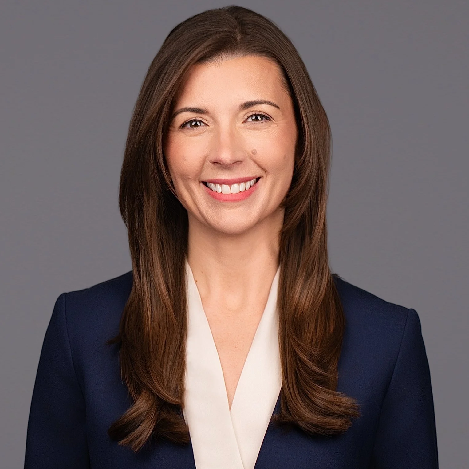 Headshot of Melody Wells with long brown hair, wearing a navy blazer and cream blouse, smiling against a gray background.