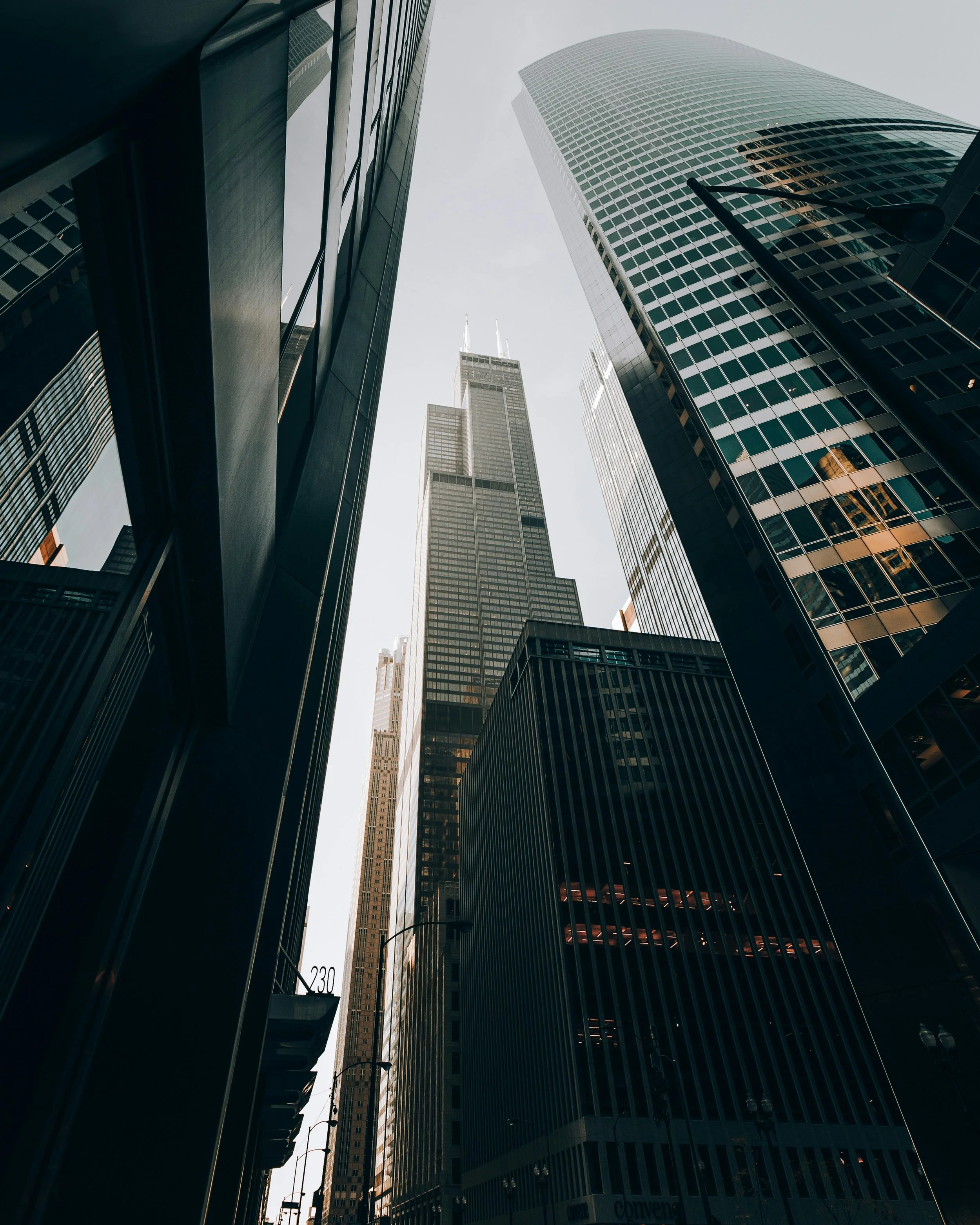 View of tall modern skyscrapers from street level in a city, looking upward, with a cloudy sky.