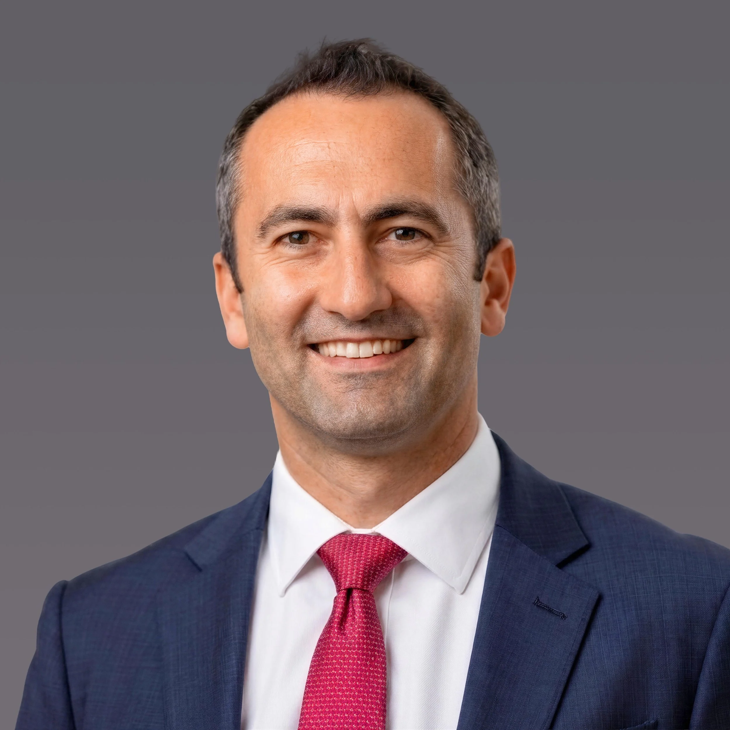 Headshot of a Matthew Jacobs smiling in a navy suit, white shirt, and red tie against a gray background.