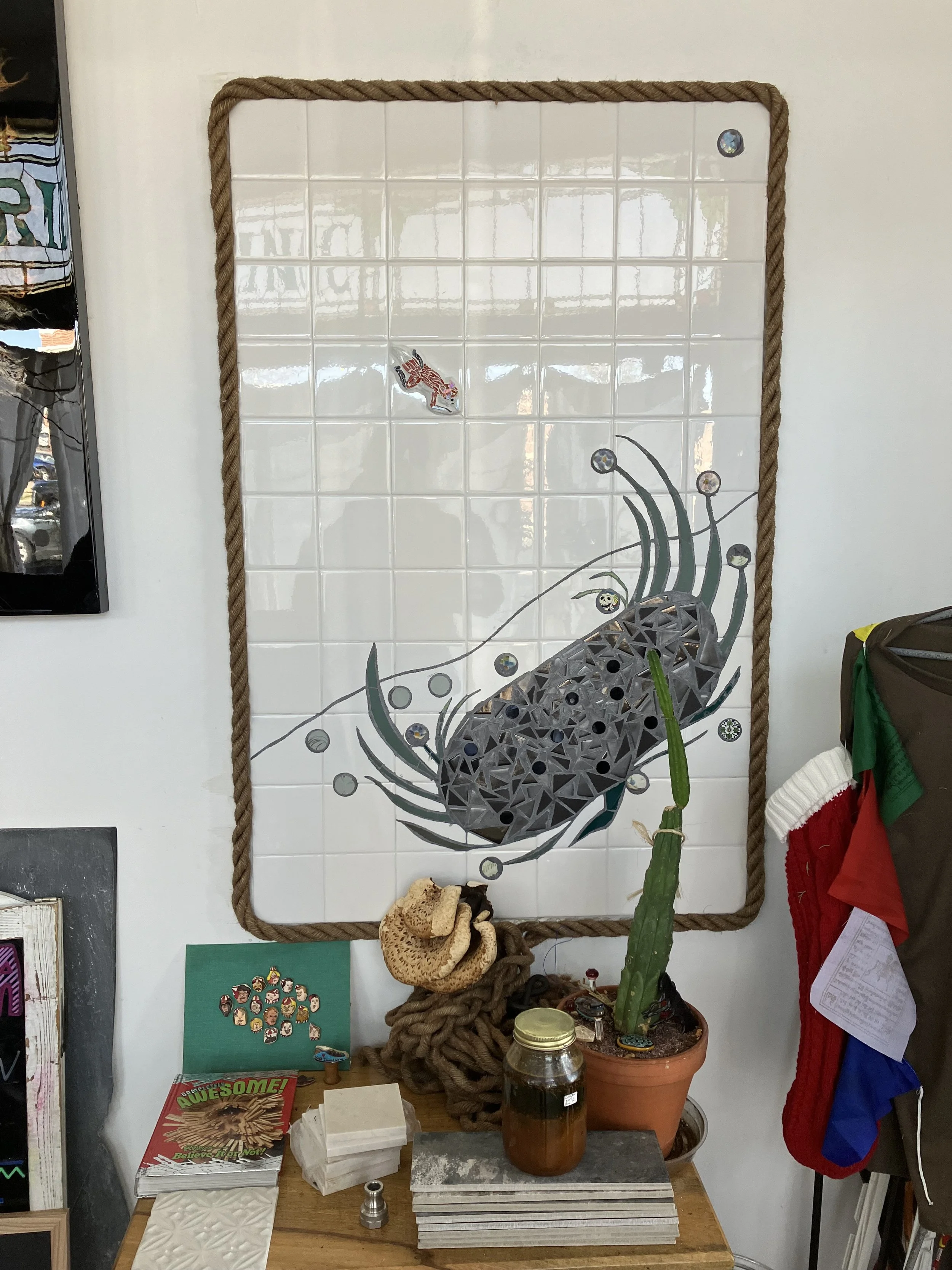 Decorative wall art featuring a mosaic of a lobster on a white tiled board framed with a thick rope. A cactus in a pot and various small objects sit on a wooden table below the mosaic.