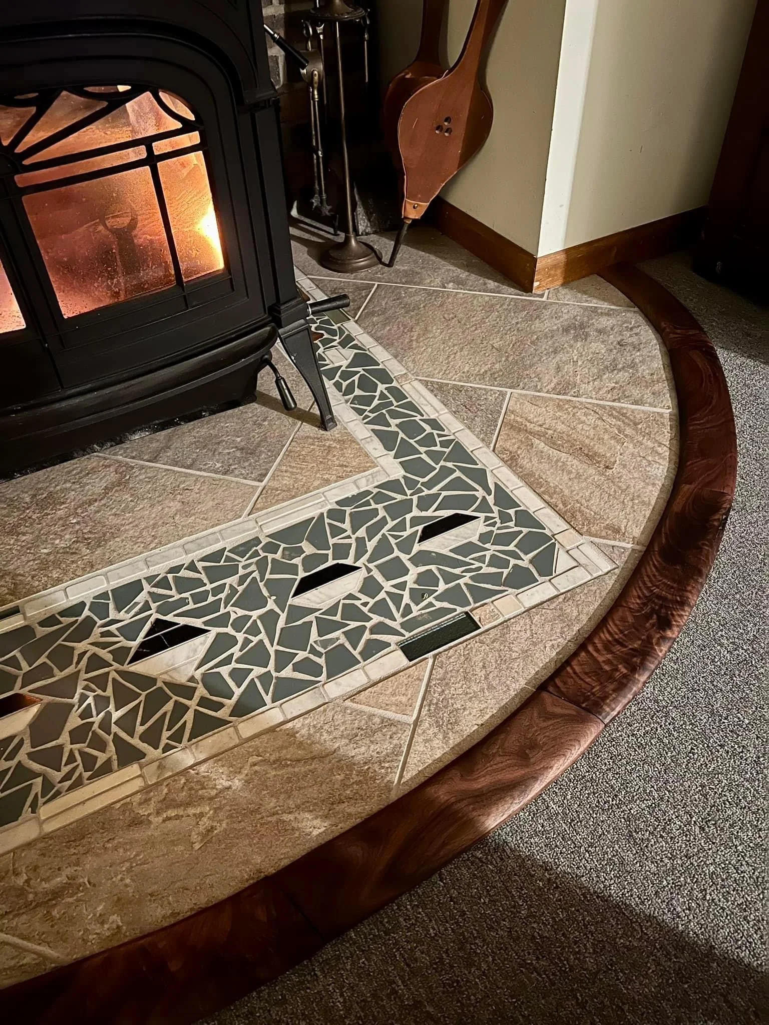 Close-up of a fireplace hearth featuring a stone and tile mosaic design, surrounded by a wooden border, and a portion of a black wood stove with a fire burning inside.