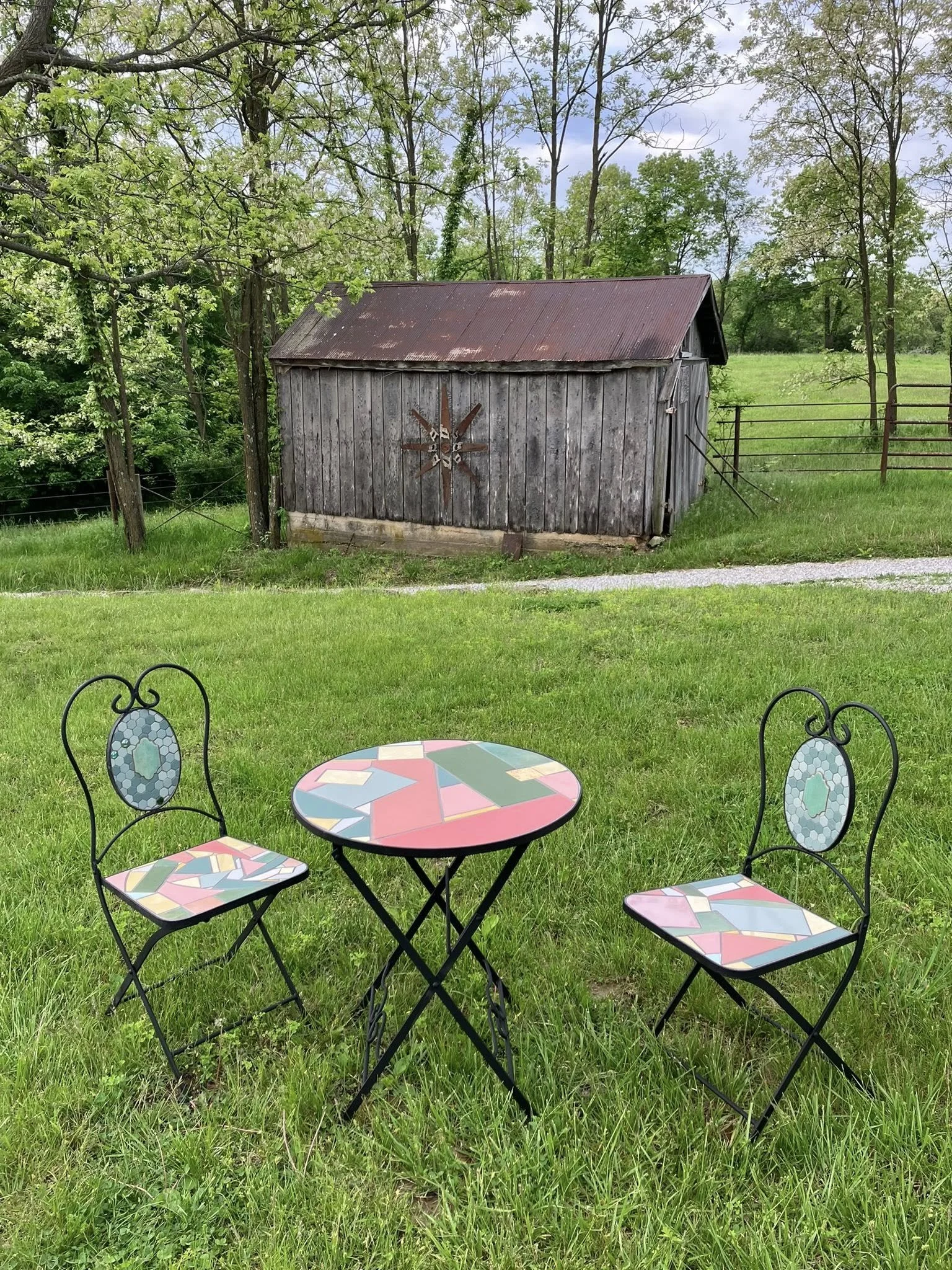 Colorful mosaic table with two matching mosaic chairs on green grass, old wooden shed with rusted metal roof and decorative metal star in the background, trees and overcast sky.