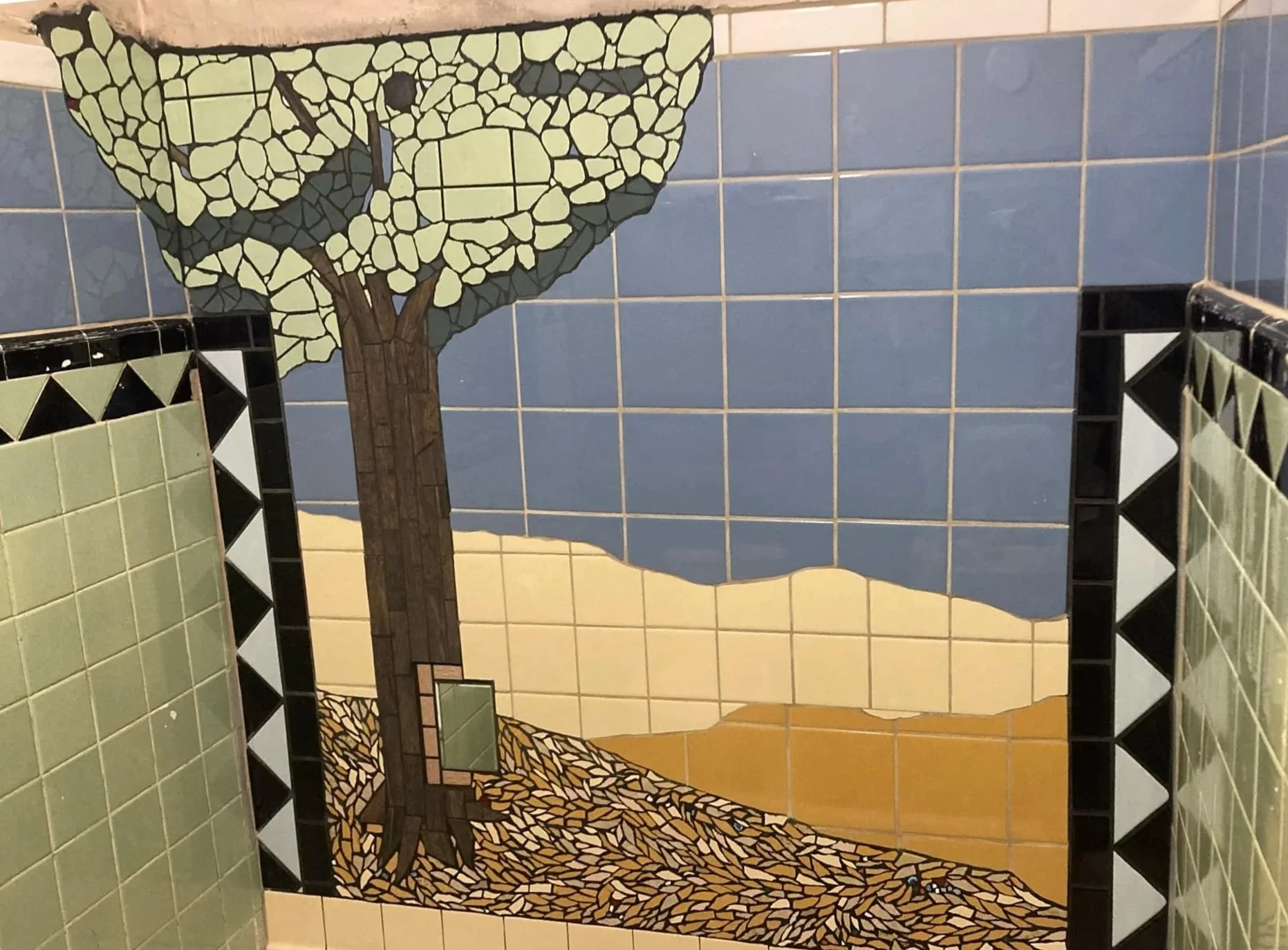 Mosaic mural of a tree, sky, and ground, located between two tiled walls.
