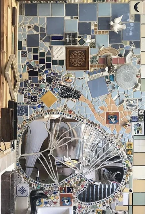 Wall decorated with various colorful mosaic tiles, mirrors, and small decorative objects, including a large bicycle-shaped mirror and art pieces