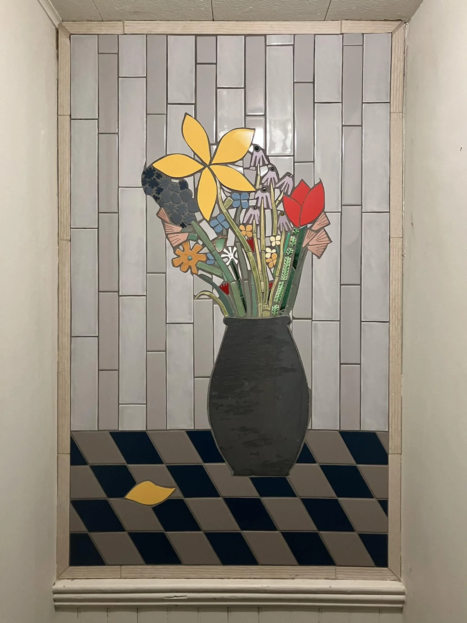 Colorful mosaic of a black vase with a bouquet of flowers, including yellow, red, purple, orange, and blue, set against a white tiled background and a checkered floor.