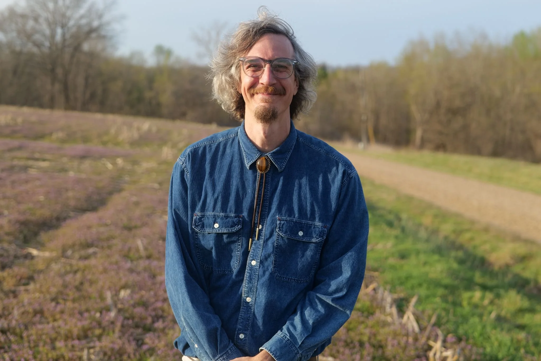 A man with glasses and long gray hair standing outdoors in a field with purple flowers and trees in the background.