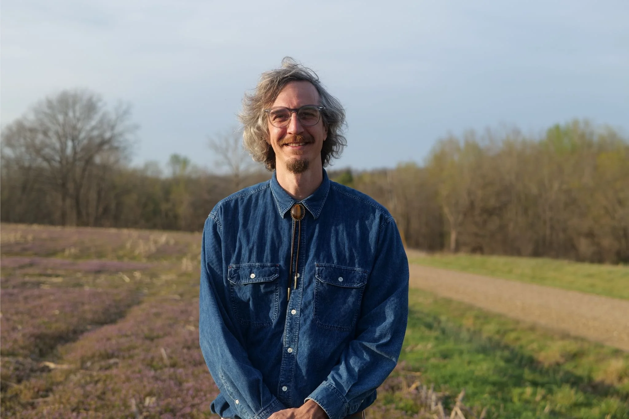 A man with glasses, gray hair, and a beard stands outdoors in a field of purple flowers, smiling at the camera.