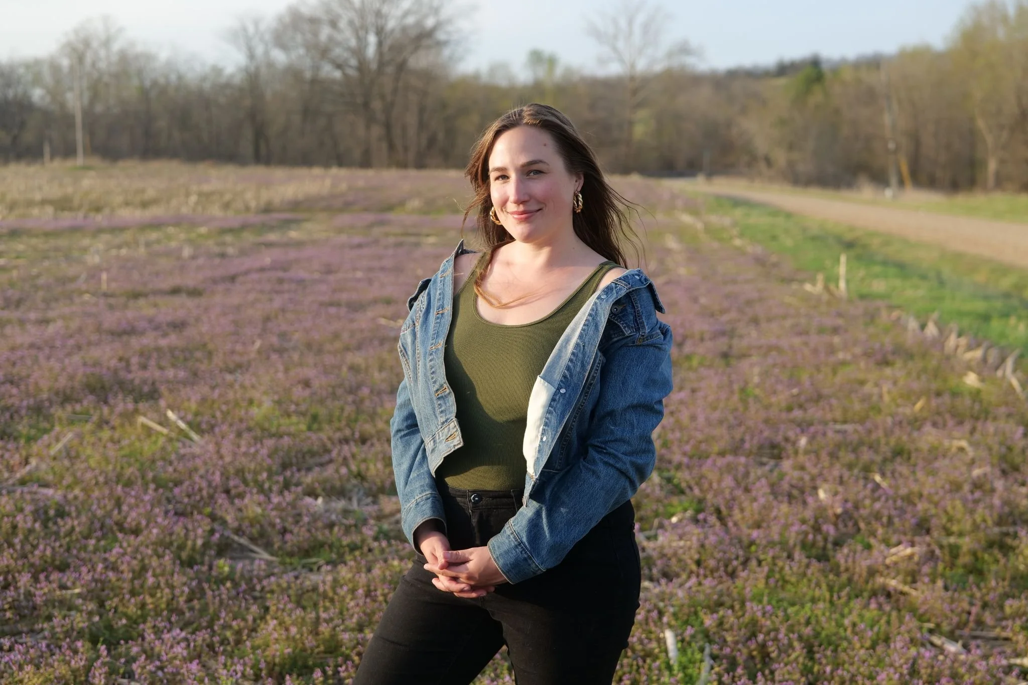 A woman standing outdoors in a field of purple flowers with trees in the background, wearing a denim jacket over a green top and black pants, smiling at the camera.
