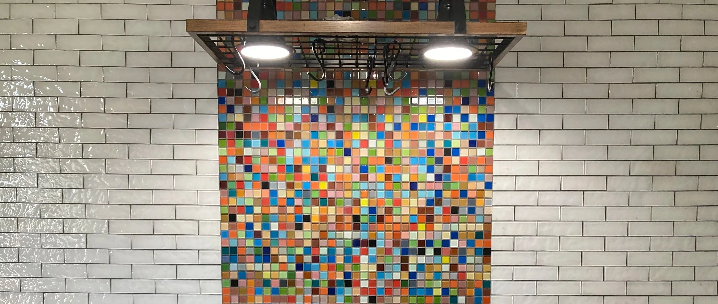 A mosaic wall with small colorful tiles arranged in a random pattern, framed by gray brick walls on both sides and overhead metal and wood shelving with two bright ceiling lights.