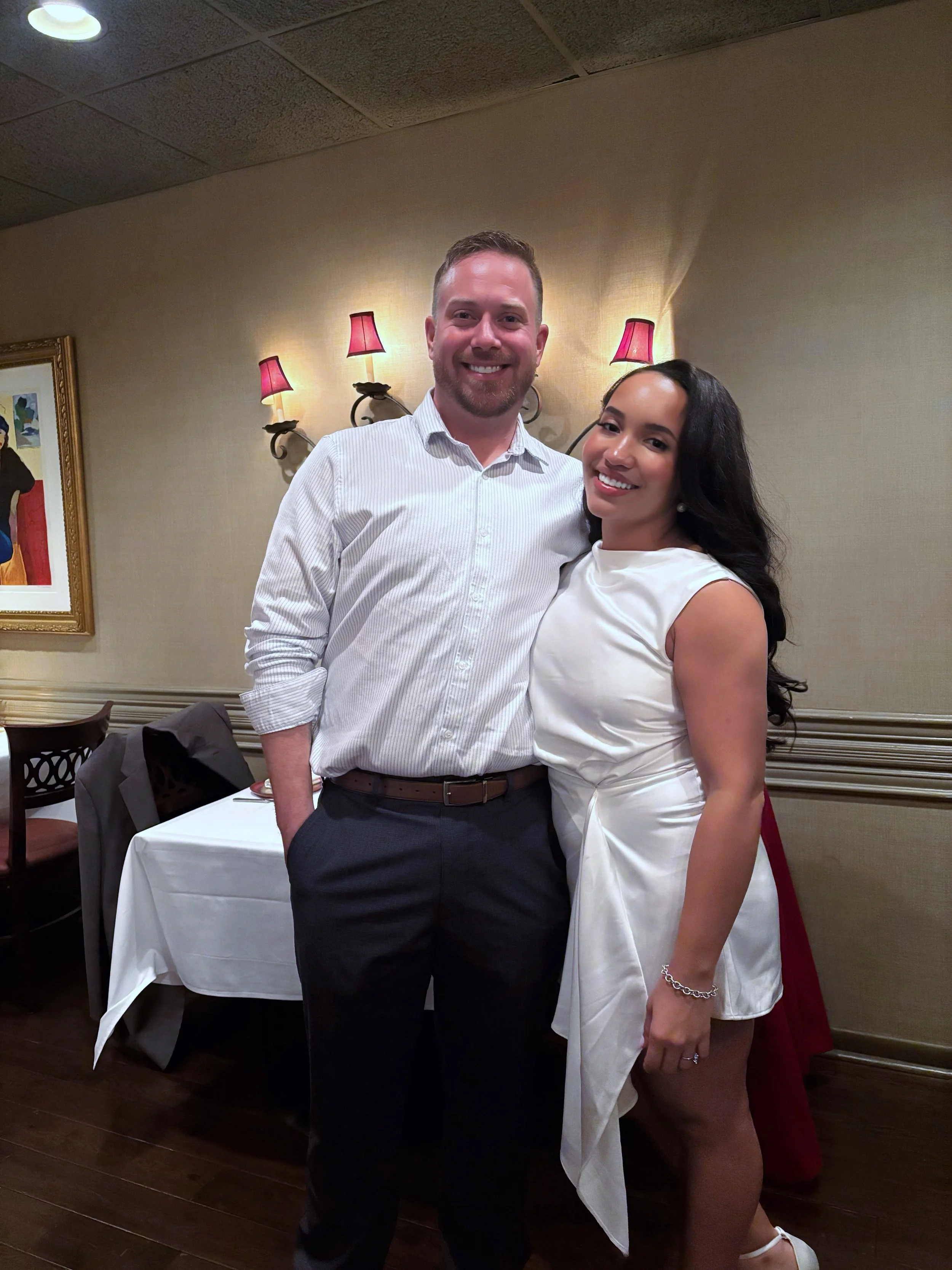 A man and woman smiling and posing together in a restaurant or banquet hall. The man is wearing a white dress shirt and dark pants, and the woman is wearing a white dress with a bracelet. They are standing close to each other, with warm lighting and framed artwork on the wall behind them.