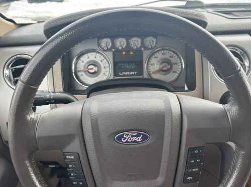 Close-up of a Ford F-150 Lariat steering wheel and instrument cluster with white-faced gauges and the Ford logo.