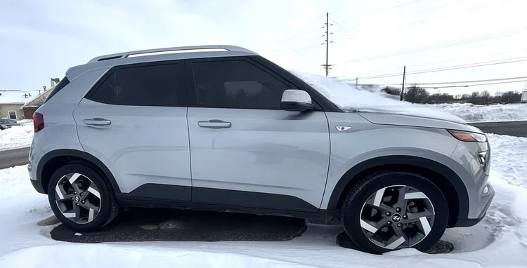 Passenger side profile of a silver Hyundai Venue parked in a snowy parking lot with snow on the roof and windshield.