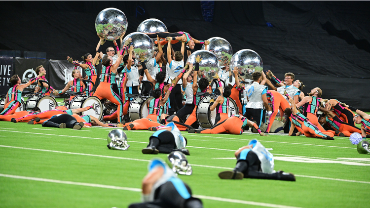 Performers in vibrant orange and teal uniforms pose with large reflective silver spheres and marching drums during a field performance.
