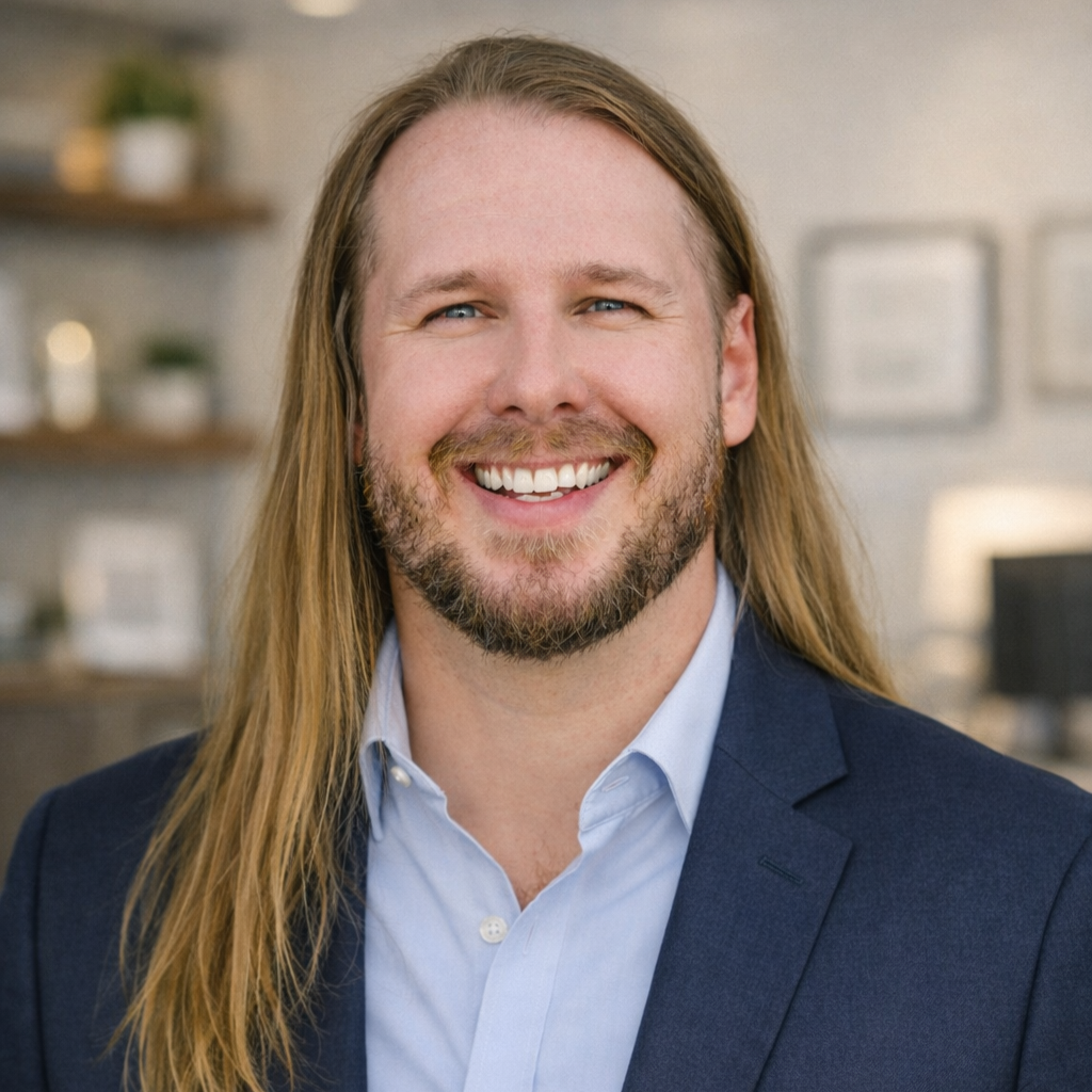 A man with long blond hair, a beard, and a bright smile wearing a blue blazer and a light blue dress shirt inside an office space.